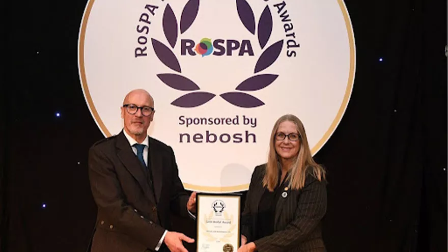 Global health and safety award winners. Two people  holding a certificate and smiling for the camera in front of The Royal Society for the Prevention of Accidents (RoSPA)  image