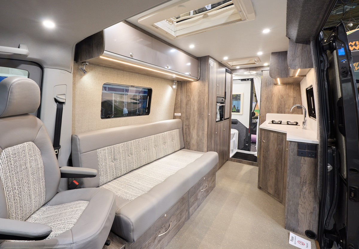 The Rolling Homes Camper Ltd Darwin FL has beige carpet.  The upholstery is mid brown with a stripped pattern.  The cabinets are pale brown with wooden surrounds.  The interior door is open revealing the rear washroom.  There are two roof lights.