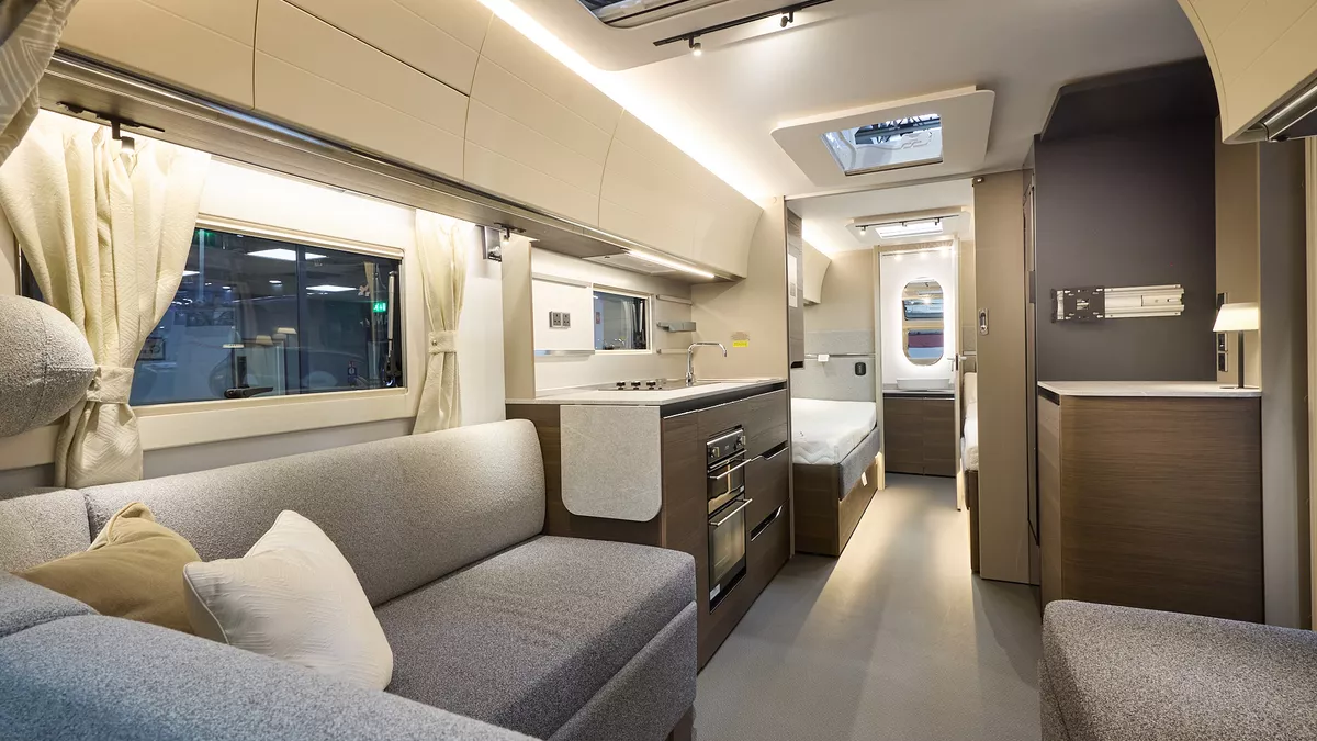 Interior of the Adria Alpina Colorado with grey sofas, cosy cushions and wood accents, illuminated by natural light through windows and skylights, offering a view outside.  Perfect for relaxation and designed for comfort.