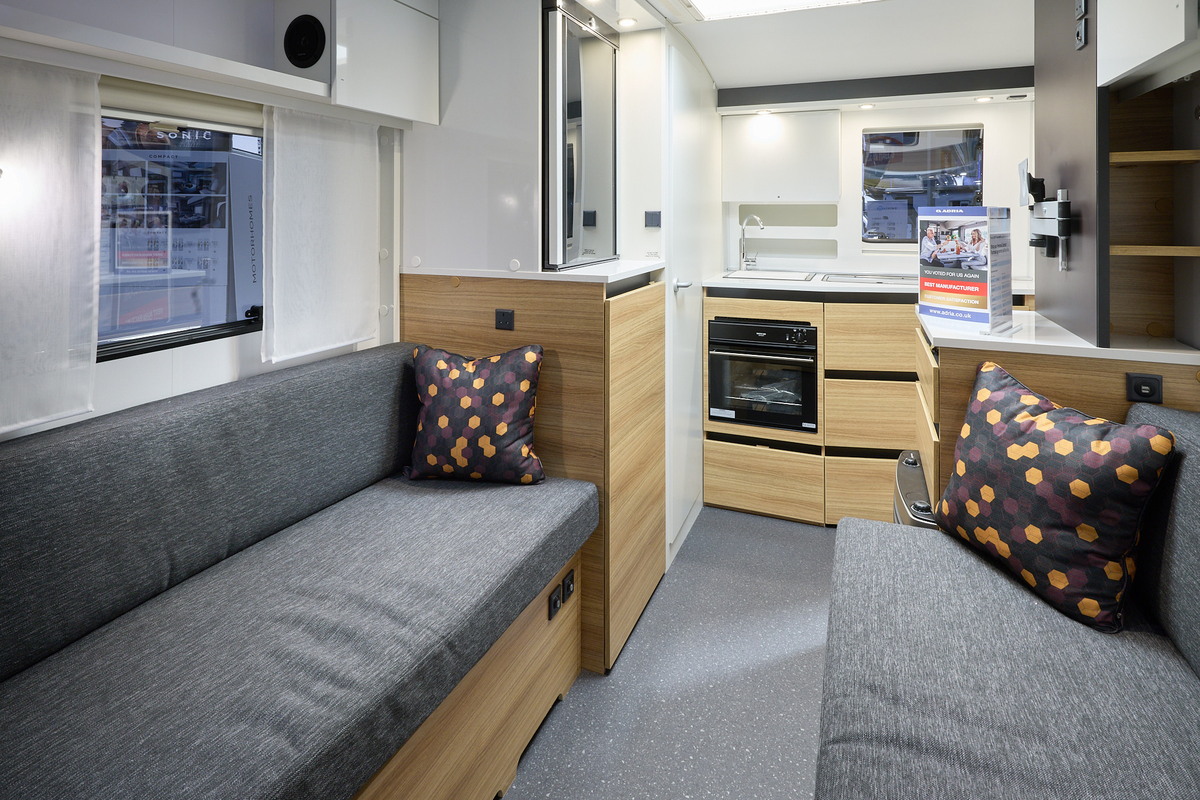 The interior of the Adria Action 361 LT has white walls.  The furniture is mid wood.  There are two sofas with grey upholstery and a cushion with a hexagon pattern on each.  The kitchen is at the rear with a full oven under the sink with a mixer tap.