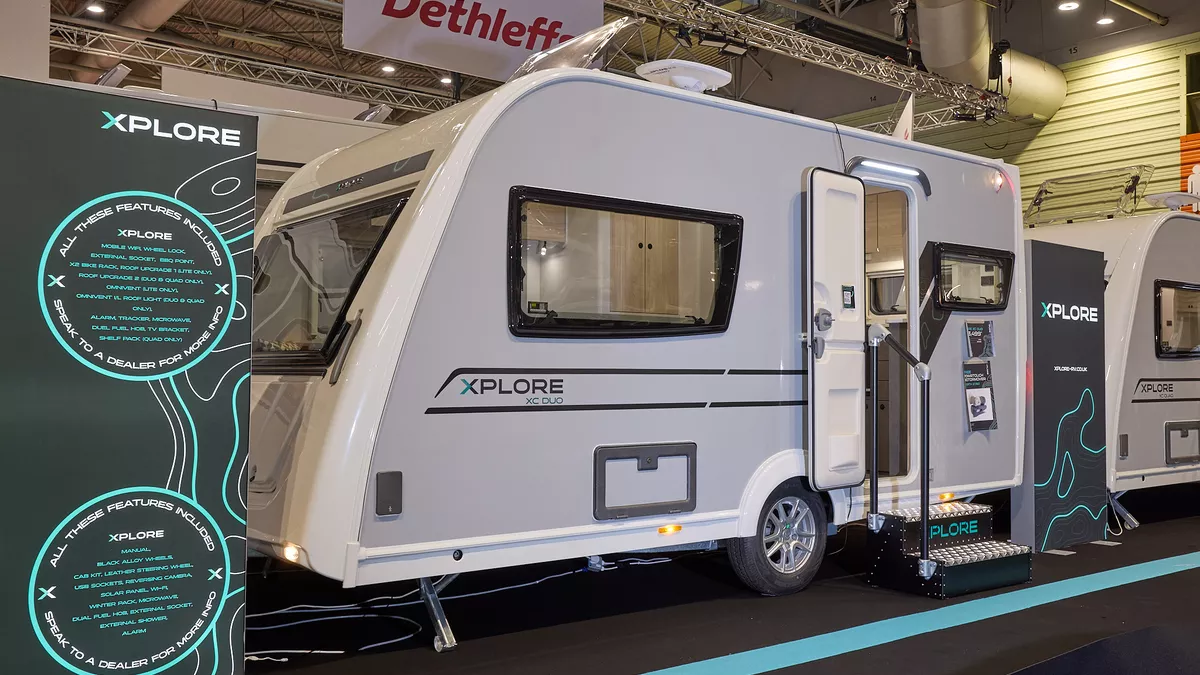The Xplore XC DUO is white with grey sides and black decals.  There are two metal steps to gain easy access.  There is a large Xplore advertising banner next to the DUO. 
