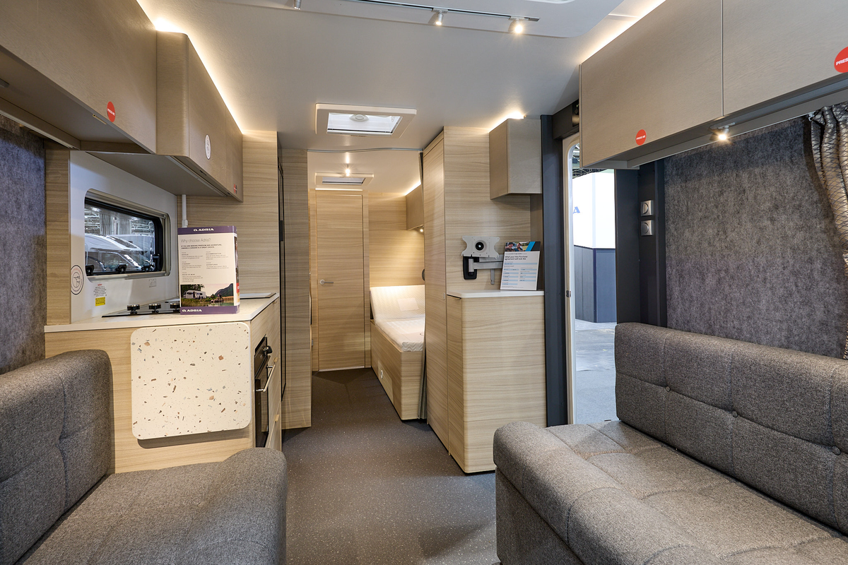 Adria Altea Tyne’s interior, it is grey with light wooden cabinets.  The upholstery is grey.  The fixed bed is beyond the kitchen.  The entrance door is open.