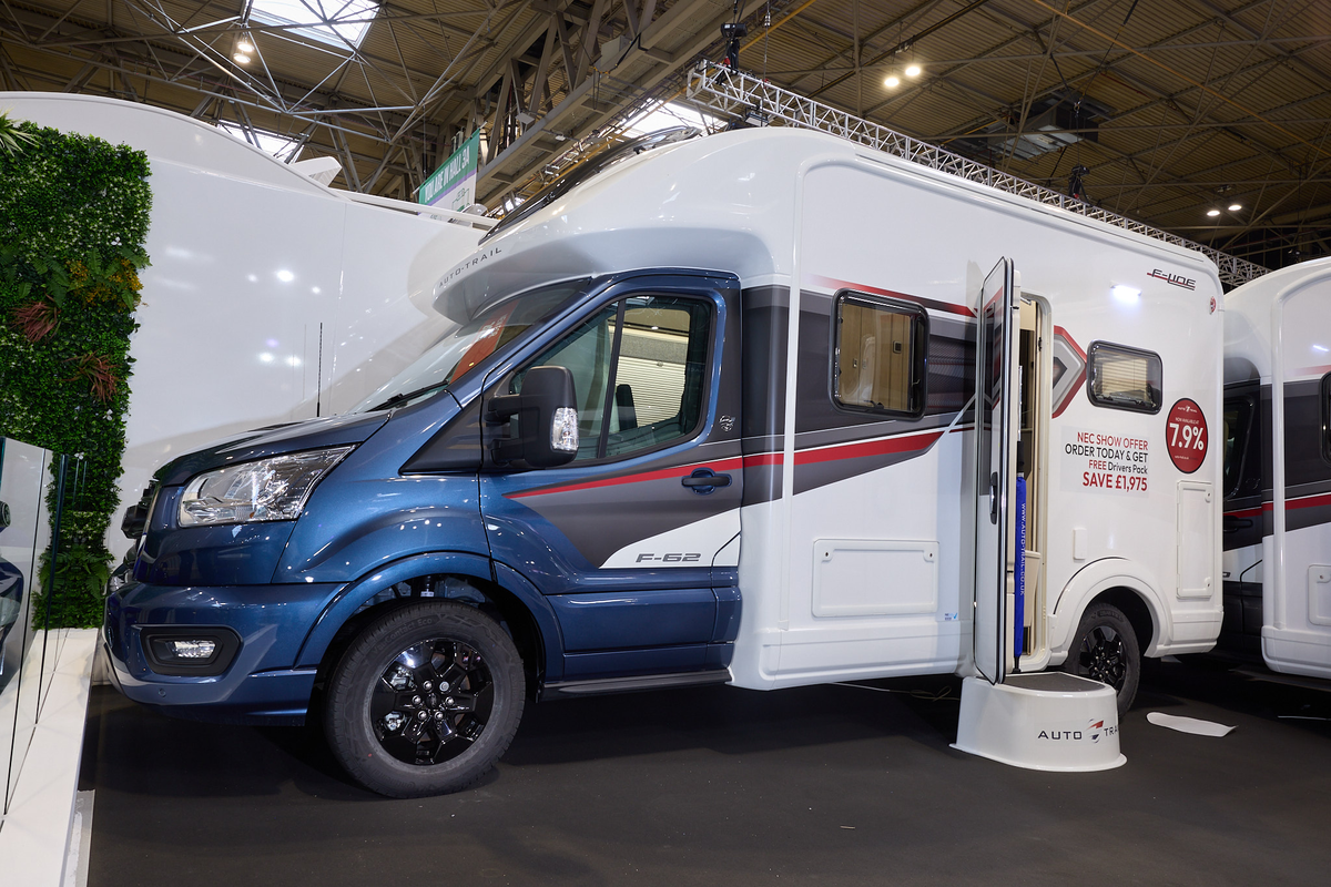 The Auto-Trail F-Line F62 has a blue cab and white body with grey and red decals.  There is a show offer on its side in red.  There is a white step by the opened habitation door.  