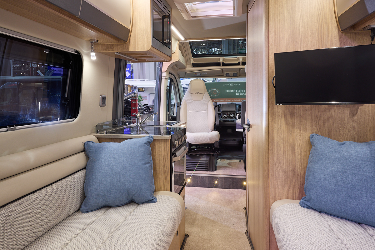 The Auto-Trail V-Line 540 SE has beige carpets, with pale wood cabinets with cream and grey overhead lockers.  The upholstery is white and cream.  There are two blue cushions.  The front seats are swivelled around.  There is a roof light.