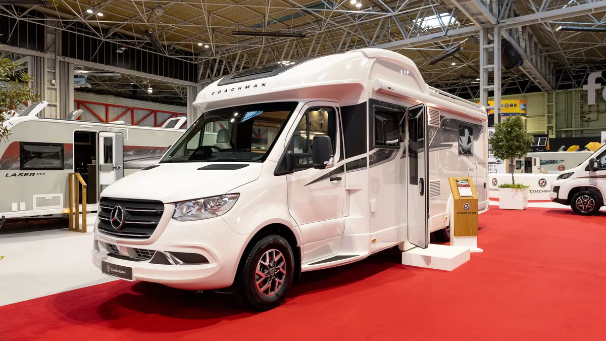 The Coachman Travel Master 545 is white, it is a Mercedes Benz.  Its habitation door is open and there is step to gain access.  Next to the step is a wooden stand