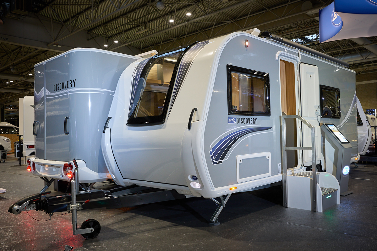 Bailey Discover D4-4 exterior’s, grey and white with a large single front window.  The entrance door is open and there are fixed metal steps into its interior.  There is an interactive stand next to the door.