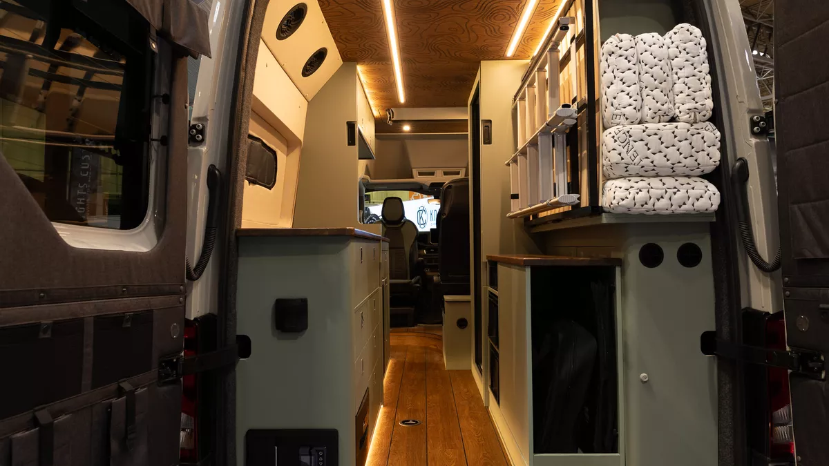 The Knights Custom Conversions Prestige XL has a wooden ceiling with a pattern, the cabinets are green with wooden edging.  The floor is wooden and there is a stowed ladder to the right.  The folded mattress is in the corner.