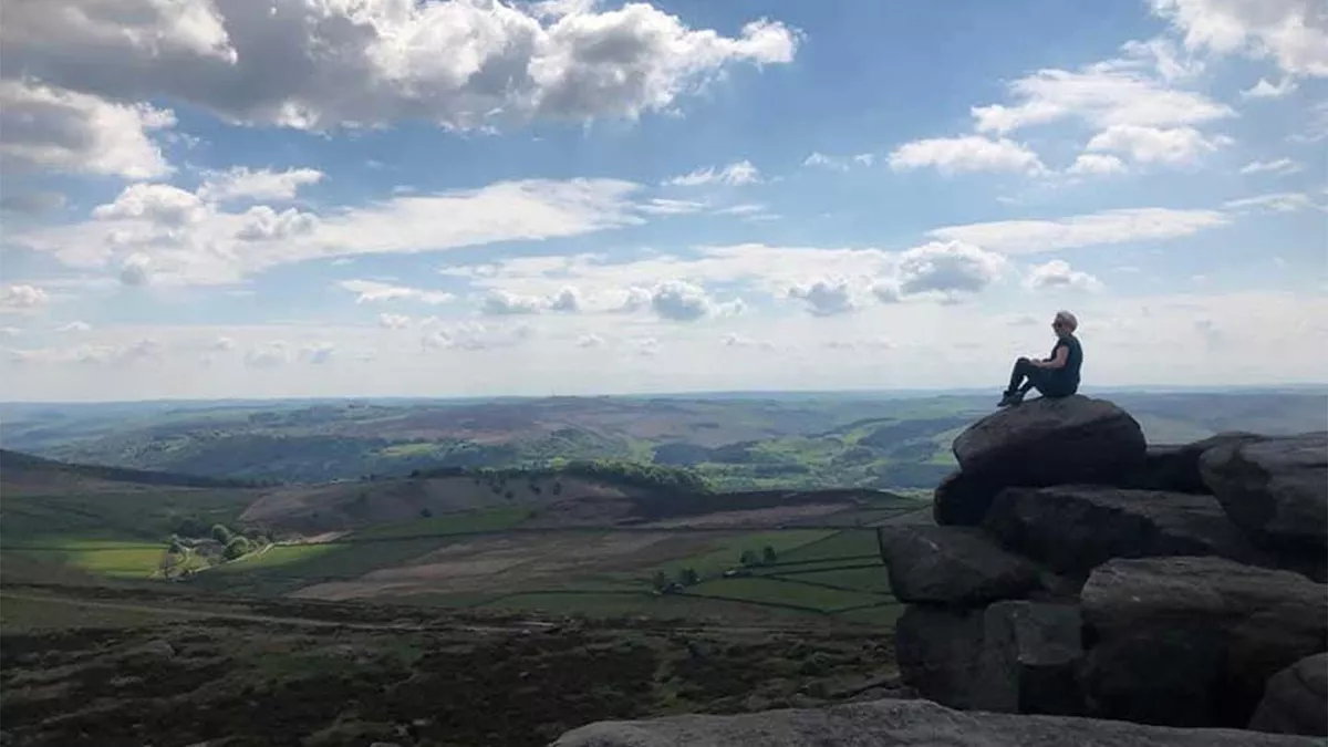 A person sitting high on a rock on the top of Stanage Edge looking out over the rolling hills that go on more miles under a blue sky of white clouds