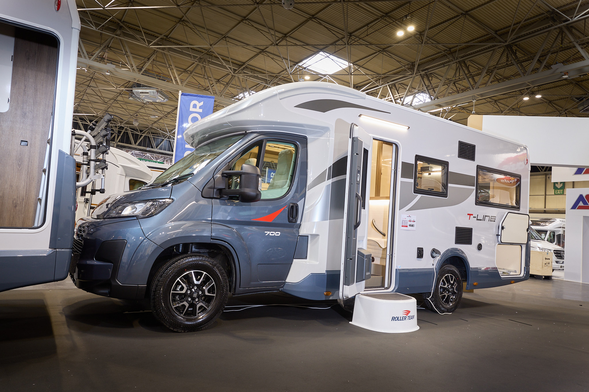The Roller Team T Line 700 has a blue cab and its body is white with blue trim with grey and red decals.  There is a white step outside the open habitation door.  The rear locker door is open.