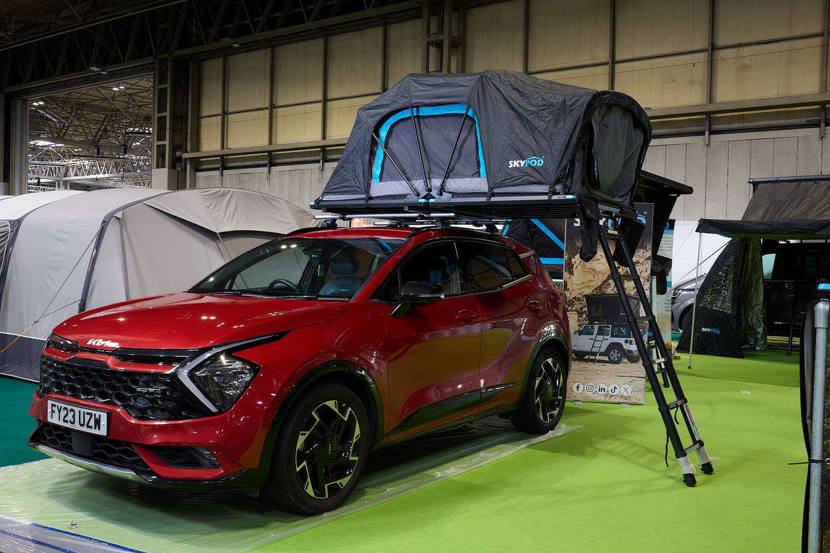 The SkyPod Lite is displayed on a Kia vehicle.  It has black canvas walls and has turquoise flashes around it.  There are three support rods by the window.  There is a black ladder.