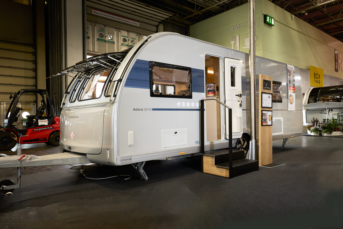 Adria Adora Seine’s exterior, white with a blue decal around the side window.  The door and front windows are open.  There are fixed wooden steps into the caravan and there’s a wooden display board to the left of the steps.   
