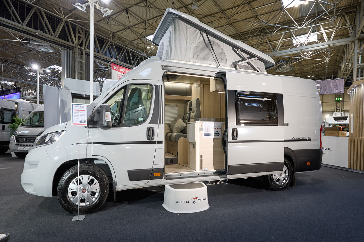 Auto-Trail Expedition 68's exterior is white with dark grey decals.  The sliding door is open revealing the lounge and kitchen.  There is a white step to gain easy access.  The rising roof is extended.