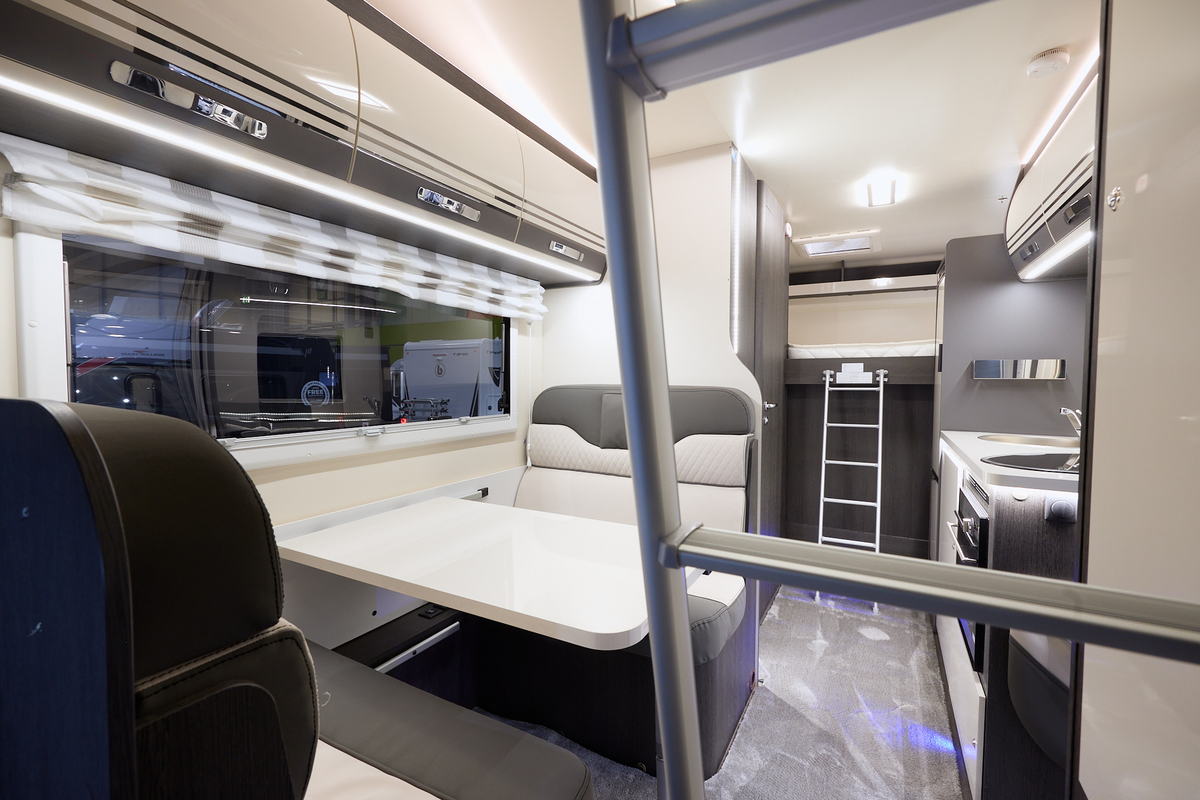 The Roller Team Zefiro 675 has grey carpet.  The upholstery is cream with dark brown highlights.  There is a cream table in front of the sofas.  The cabinets are cream with dark brown surrounds.  There are two ladders to gain access to beds.