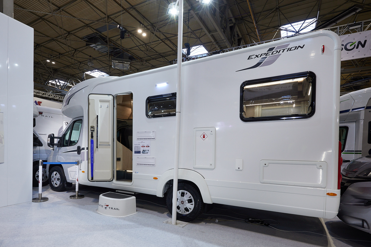 Auto-Trail Expedition C72 exterior, the motorhome is white with two windows, the habitation door is open showing into the interior, with a step to gain easy access.  There is a blue Auto-Trail umbrella in the door.