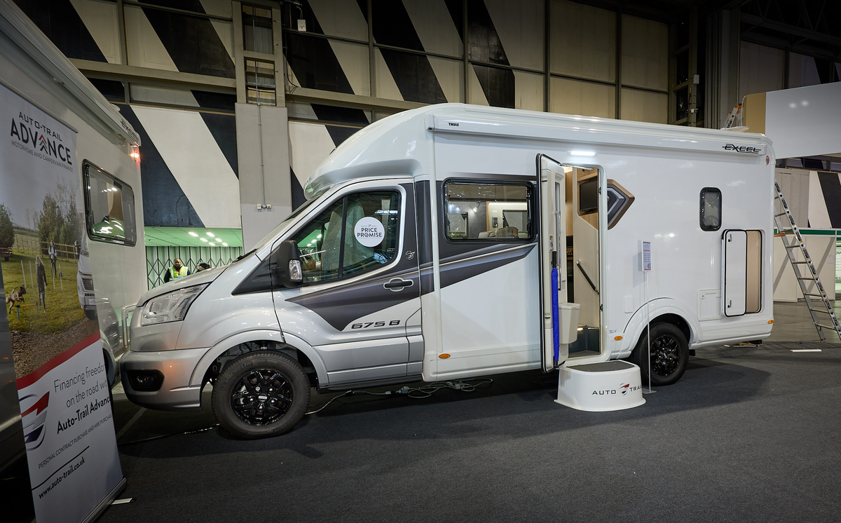 Auto-Trail Excel 675B’s exterior is white with a silver cab and dark grey decals.  Its entrance door is open revealing a handrail and a blue umbrella.  There is a step to gain easy access.