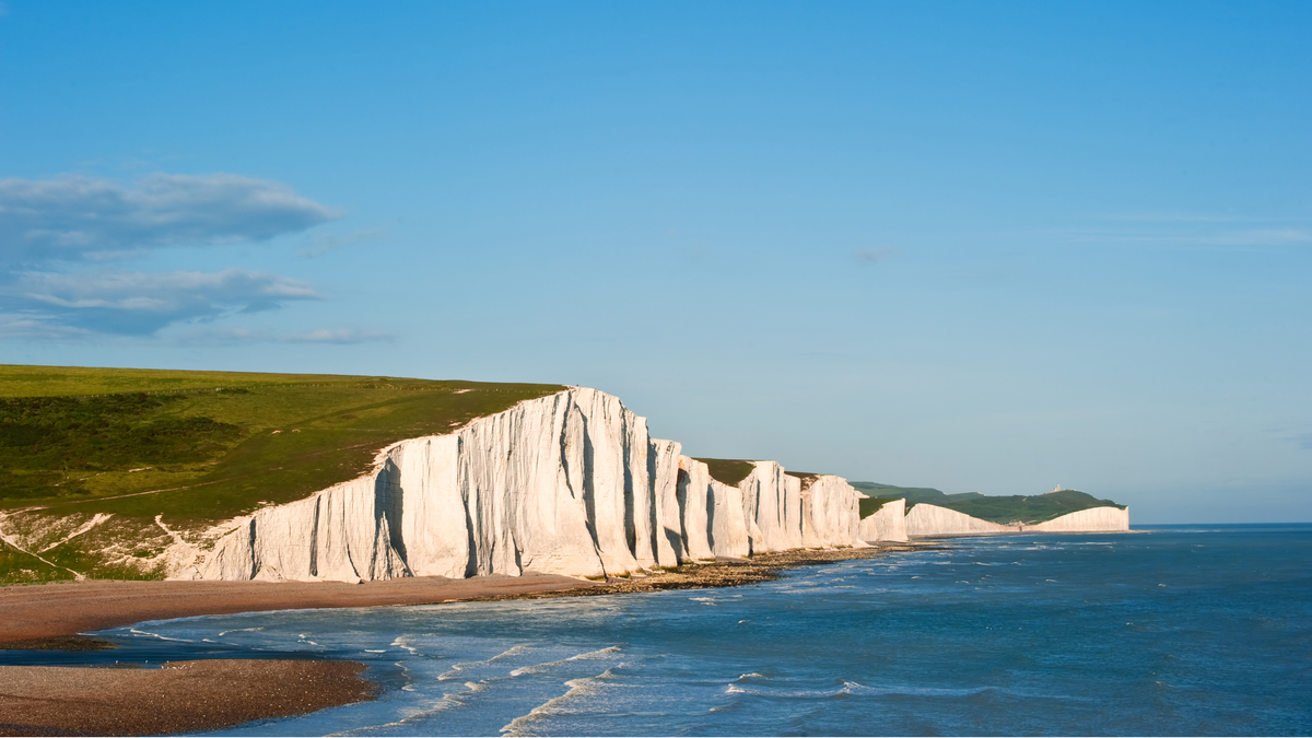 Seven Sisters cliffs in East Sussex white cliff faces topped with green grass with under a blue sky and the sea rolling onto the pebbled beach