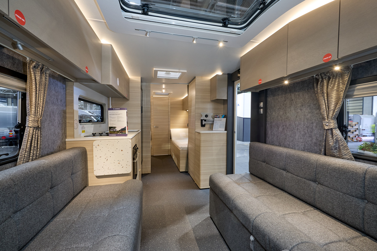 Adria Altea Tyne’s interior, it is grey with light wooden cabinets.  The upholstery is grey.  The fixed bed is beyond the kitchen.  There are three skylights.