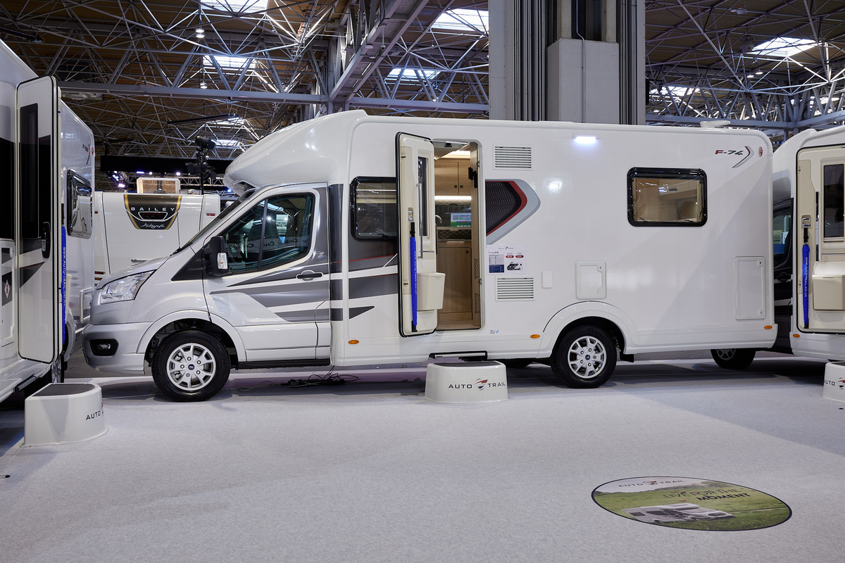 Auto-Trail F-Line F74 exterior, the motorhome is white with grey decals on the side, the habitation door is open showing into the interior, with a step to gain easy access.  There is a blue Auto-Trail umbrella in the door.