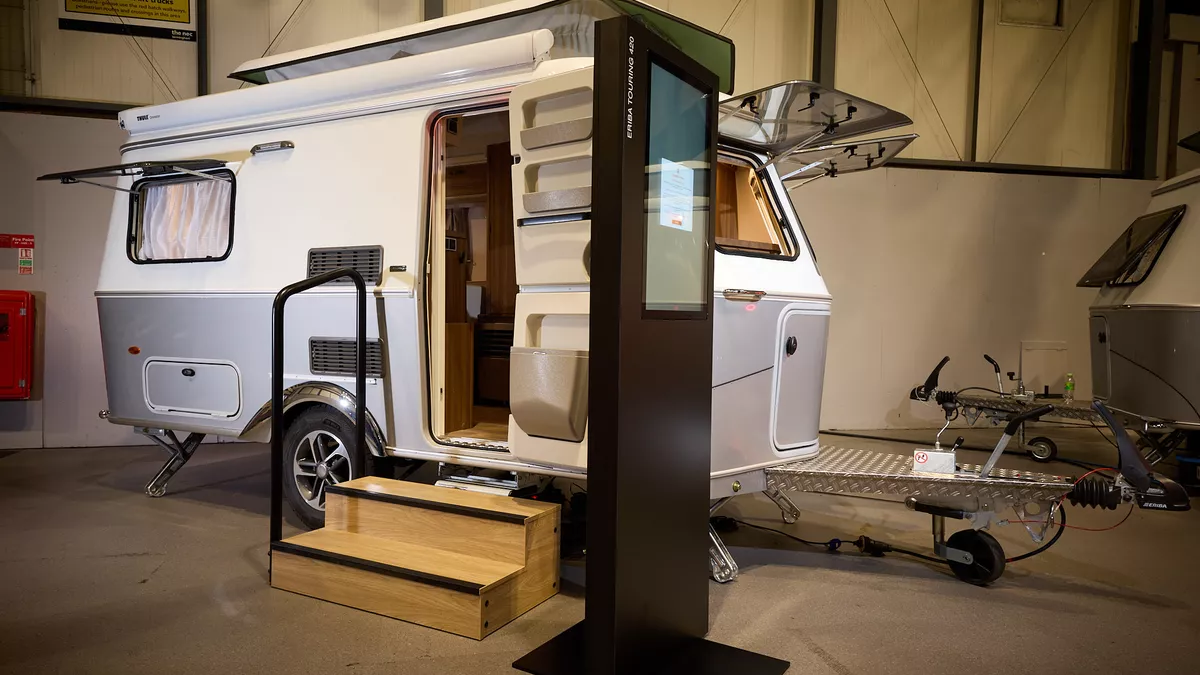 The classically styled Eriba Touring 420 Legend has a two tone layout in silver and white.  Its pop top roof is open as is its entrance door.  There are two wooden steps to gain easy access.