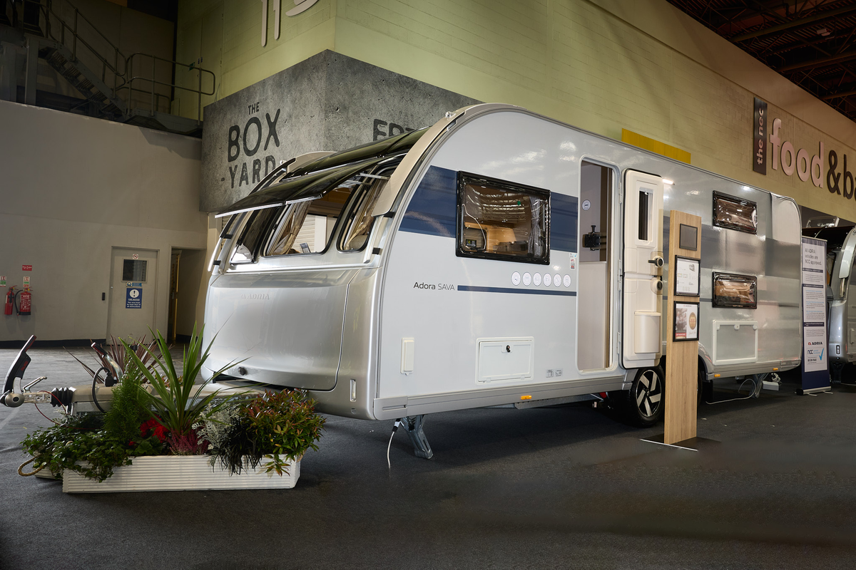 Adria Adora Sava’s exterior, white with a blue decal around the side window.  The door and front windows are open.  There are flowers in white plants around its hitch.