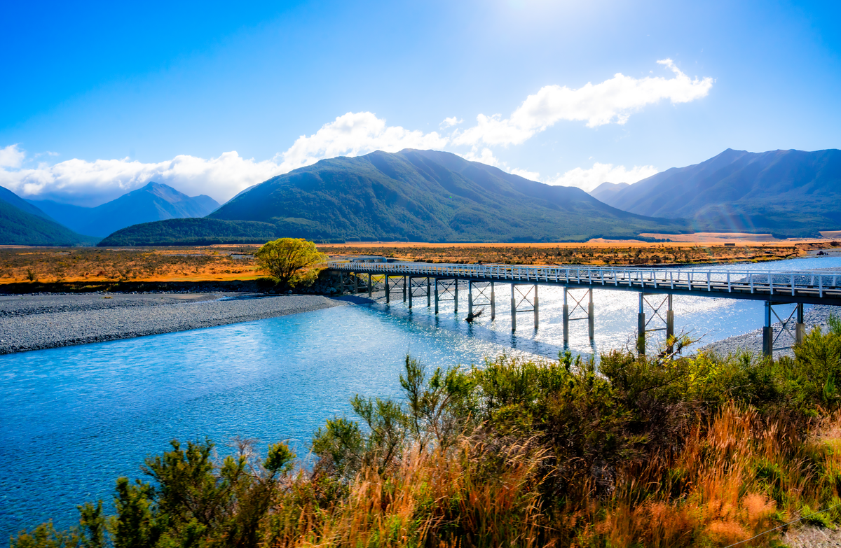 Bridge crossing over a calm river with huge mountains in the distance topped with fluffy clouds under a sunny blue sky