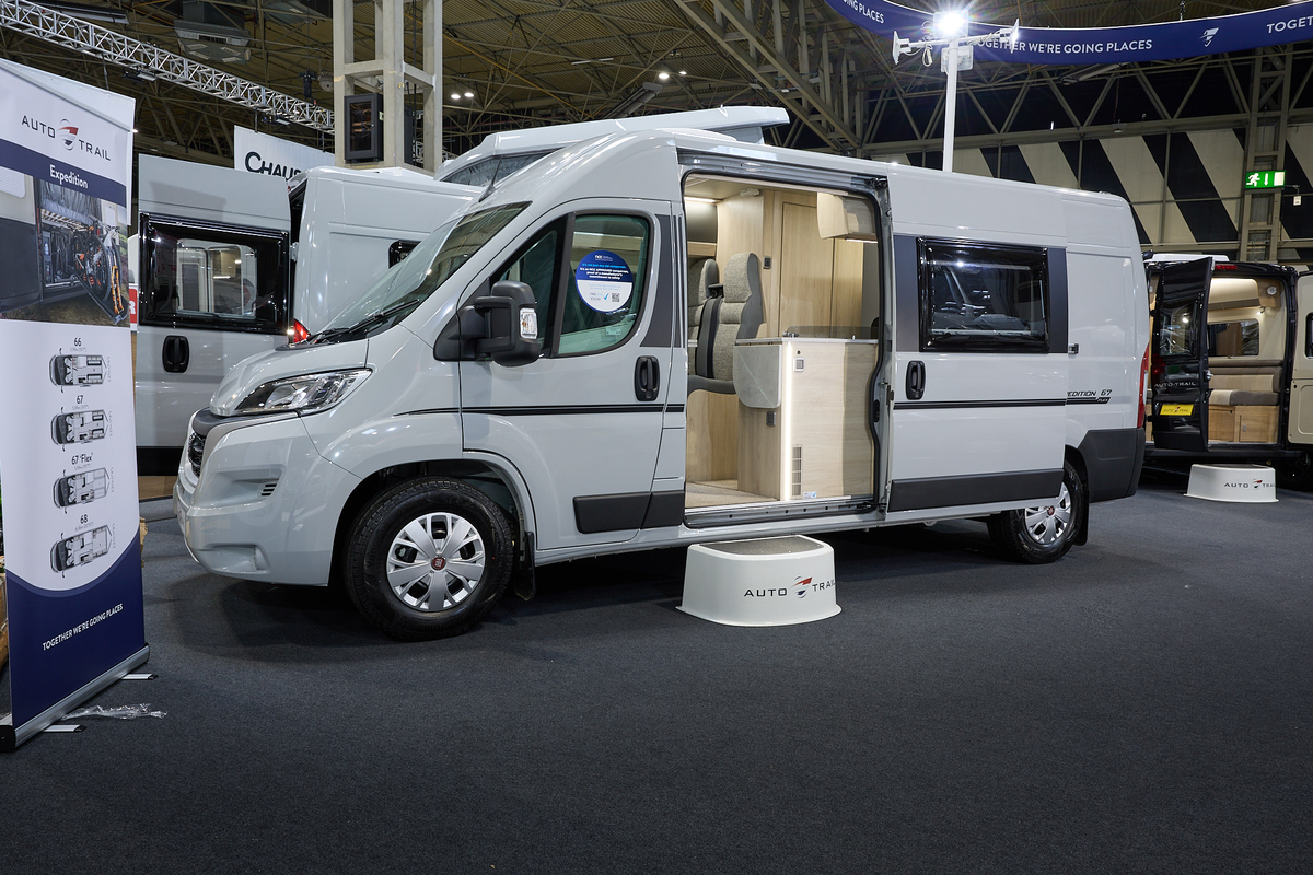 Auto-Trail Expedition 67 Flex's exterior is white with dark grey decals.  The sliding door is open revealing the lounge and kitchen.  There is a white step to gain easy access.