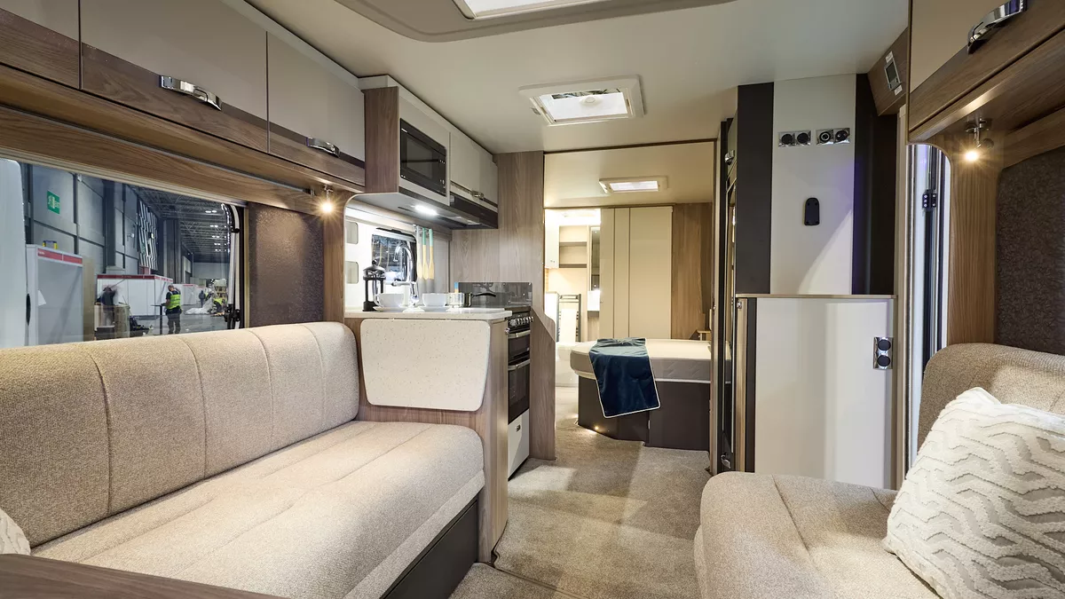 Interior of the Swift Conqueror Grande 645 with beige sofas, cosy cushions and beige carpets.   The large windows and three sky lights will let natural light flood in.  The door to the rear washroom, just beyond the bedroom, is open.