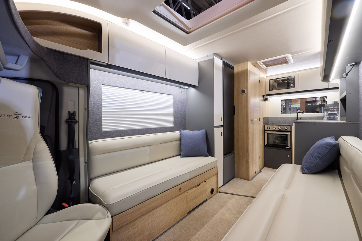 The Auto-Trail Excel 620S has beige carpet, with pale wood cabinets with white overhead lockers.  The upholstery is cream leather.  There are blue cushions.  The kitchen is at the end with an oven and a microwave in an overhead locker.