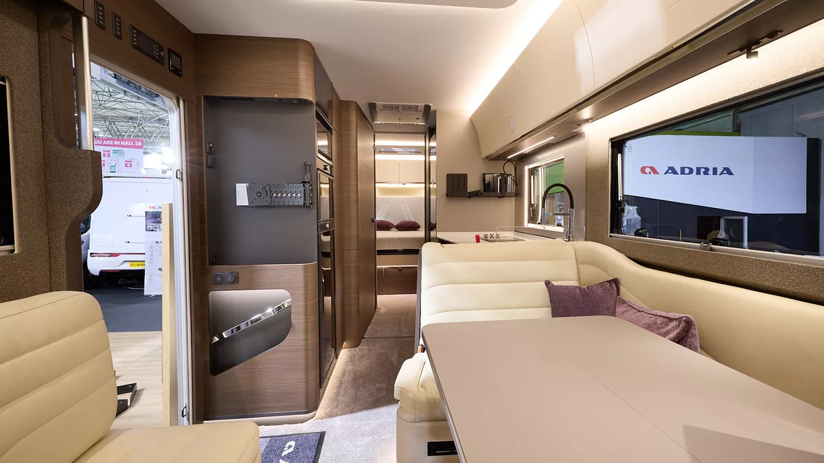 Adria Supersonic's interior has cream upholstery with curved dark wooden units.  There are two dark pink cushions, it has a sky light and a beige table.  The fixed bed is to the rear and there is a place for a TV on the left hand side.