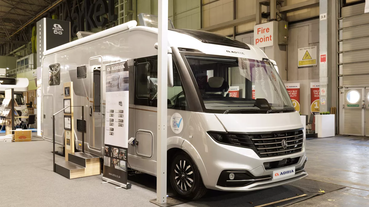 The Adria Supersonic 780 DL is silver.  There are two steps beside its habitation door.  There is a roller banner one side of the steps with an interaction stand on the other side.  Its skylights are open.