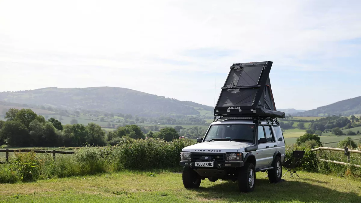 A view of a pitch at the Cider Site Certificated Location in Hereford. The grass pitch field is bordered by a fence and green bushed. A Land Rover is parked at an angle facing out. A roof top tent is open on the top of the vehicle. With views of the trees and hills in the background under a sunny but slightly hazy sky.