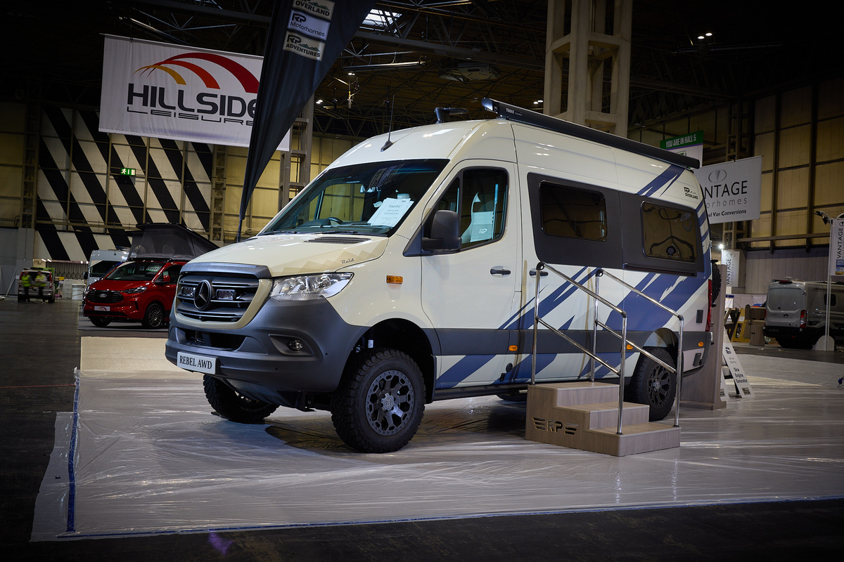 The RP Overland Rebel Raid AWD has a cream exterior with striking blue decals.  There are three steps leading to the closed sliding door.