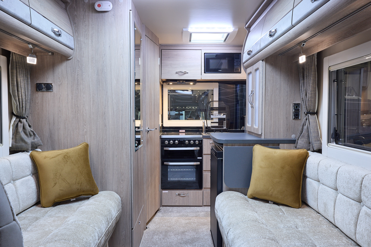 The Auto-Sleeper Nuevo has cream carpet, with pale wood cabinets with pale grey highlights.  The upholstery is cream.  There are two gold cushions.  The kitchen is at the rear with an oven and the sink cover is open.