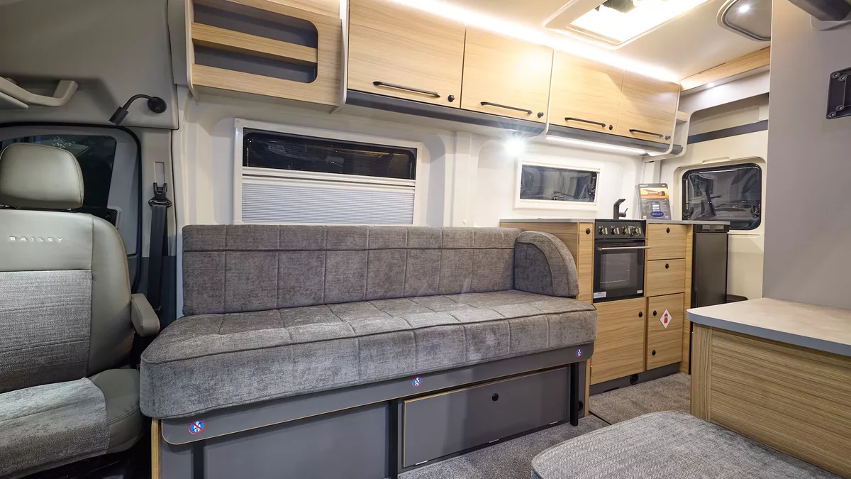 The Bailey Endeavour B63 has grey carpets, with wooden and grey cabinets.  The upholstery is also grey.  There is a kitchen to the rear of the van.  