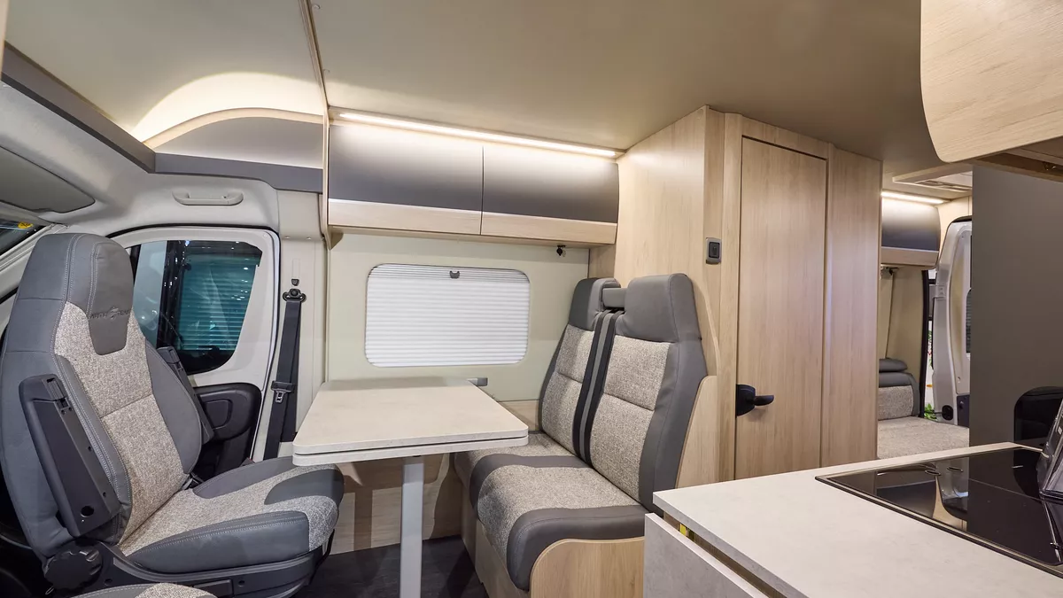 The Auto-Trail Expedition 67 has grey flooring, with pale wood cabinets with pale wood and grey overhead lockers.  The upholstery is grey and cream.  There is a rear lounge and the rear doors are open.