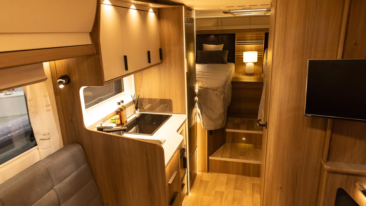 The Coachman Sportivo 565 has beige upholstery with wood furniture.  The locker doors are cream.  The kitchen is to the left.  In the rear bedroom there are fixed single beds with a beside lamp.  The TV is in the bottom right.