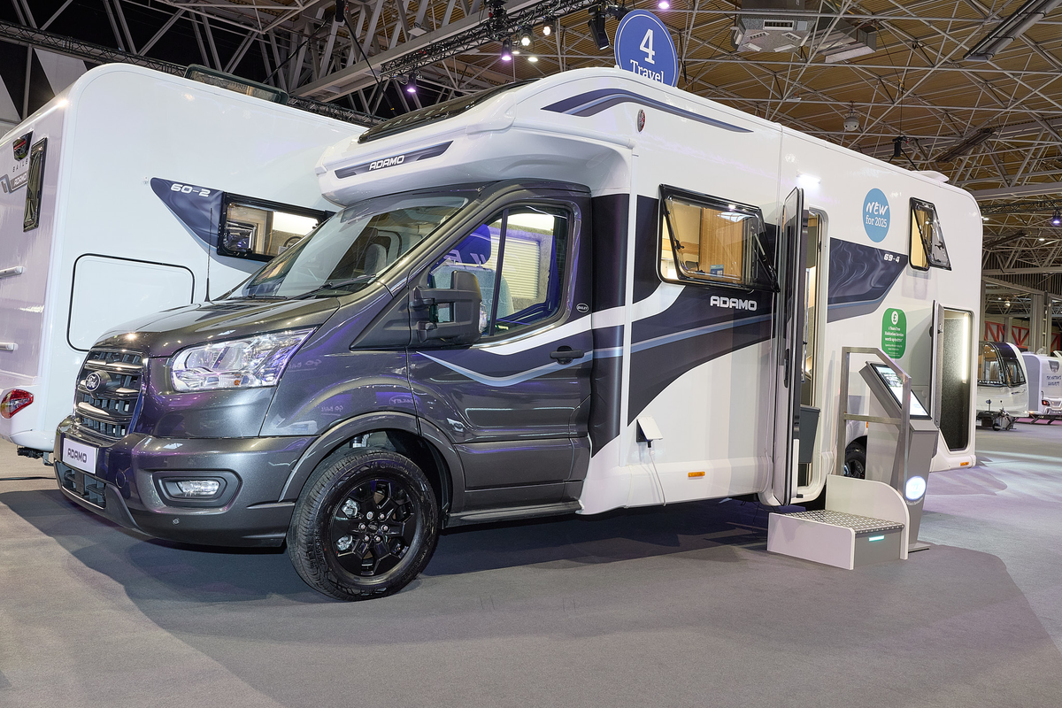 The Bailey Adamo 69-4 has a grey cab with a white body and has grey and blue decals.  There is a white and metal step by the habitation door.  There is an interactive stand to the right of the step. The rear garage door is open.