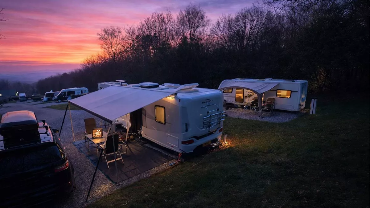 Lickpenny Matlock Club Campsite looking onto pitched caravans as the night falls with dark trees along the edge under a stunning pink, purple and orange sky as the sun sets. 