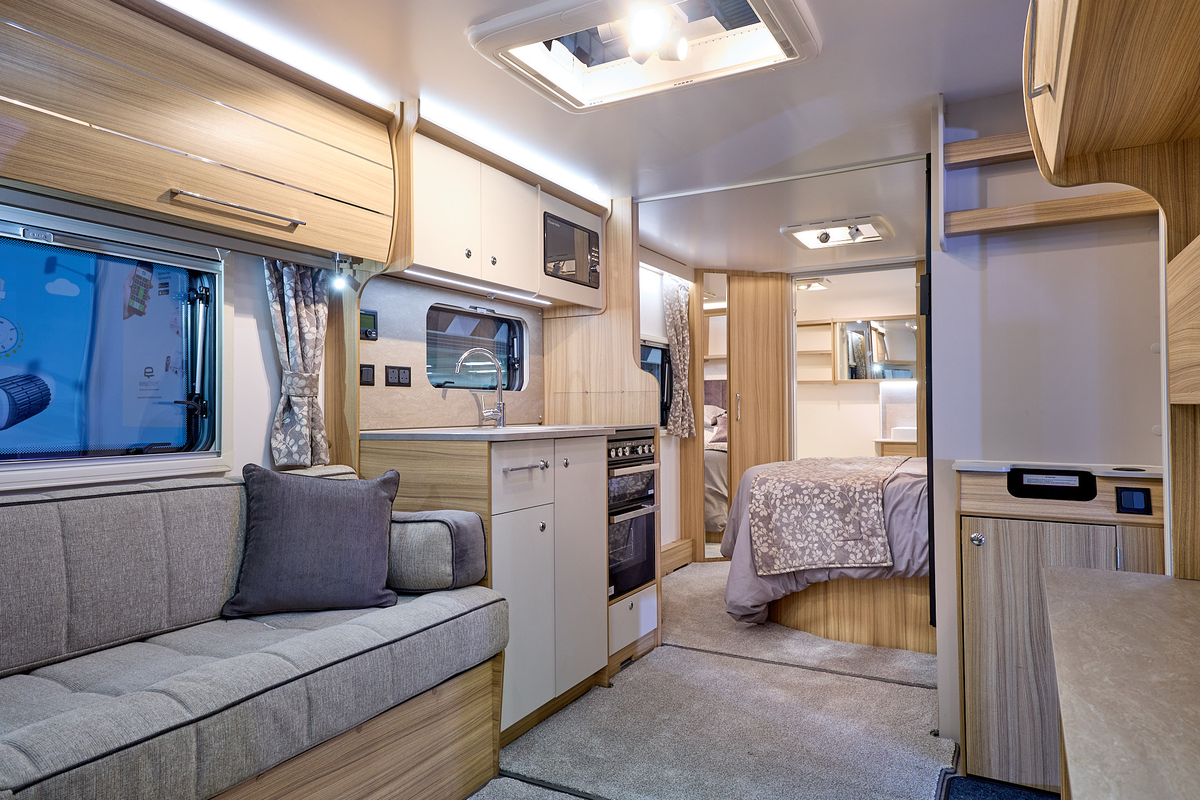 Bailey Pegasus Grande Bologna interior, it is grey with light wood.  There is a fixed bed beyond the kitchen with an end washroom, the door of which is open and is covering a large mirror.
