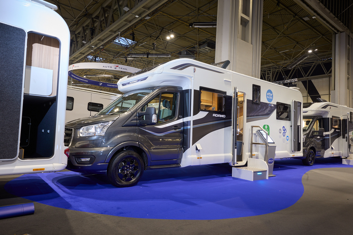 The Bailey Adamo XL DL has a grey cab with a white body and has grey and blue decals.  There is a white and metal step by the habitation door.  There is an interactive stand to the right of the step. The rear garage door is open.