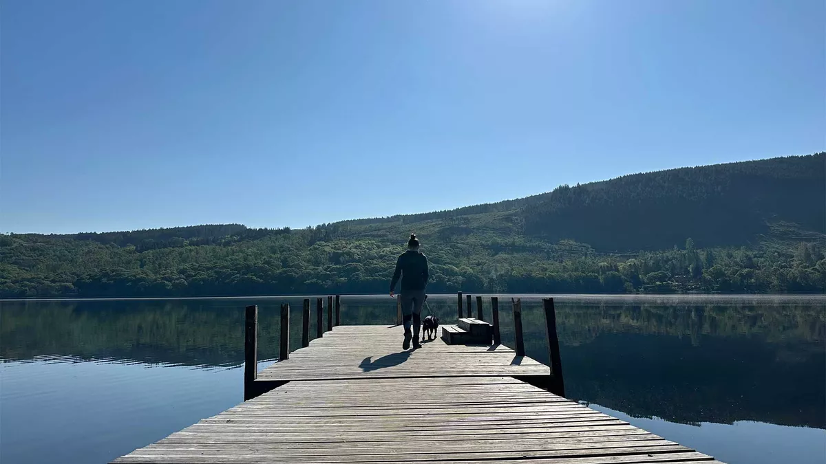 A cleat blue sky above hills and trees in the background with a person walking out onto a pier over a beautiful lake