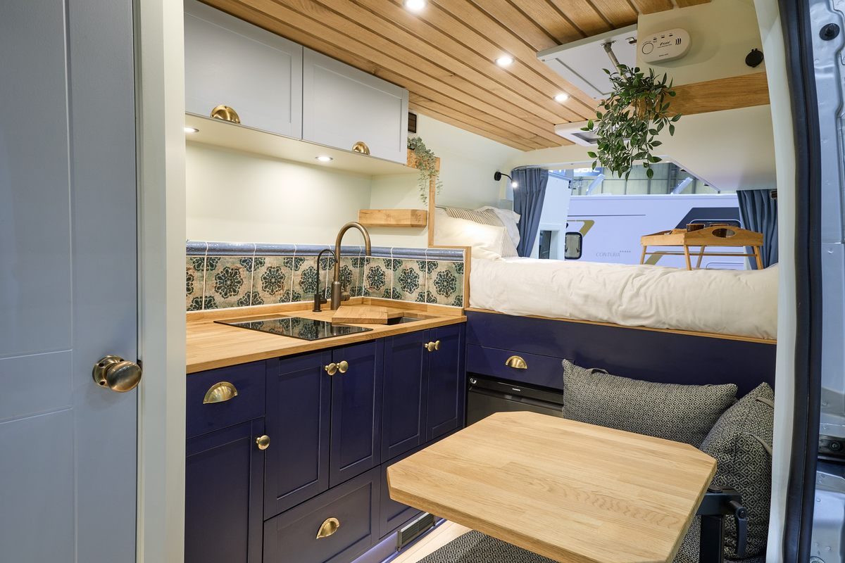 Vanlife Conversions Kunu has wooden flooring with grey upholstery, dark blue units with gold handles.  There is a fixed rear bed and the rear doors are open.