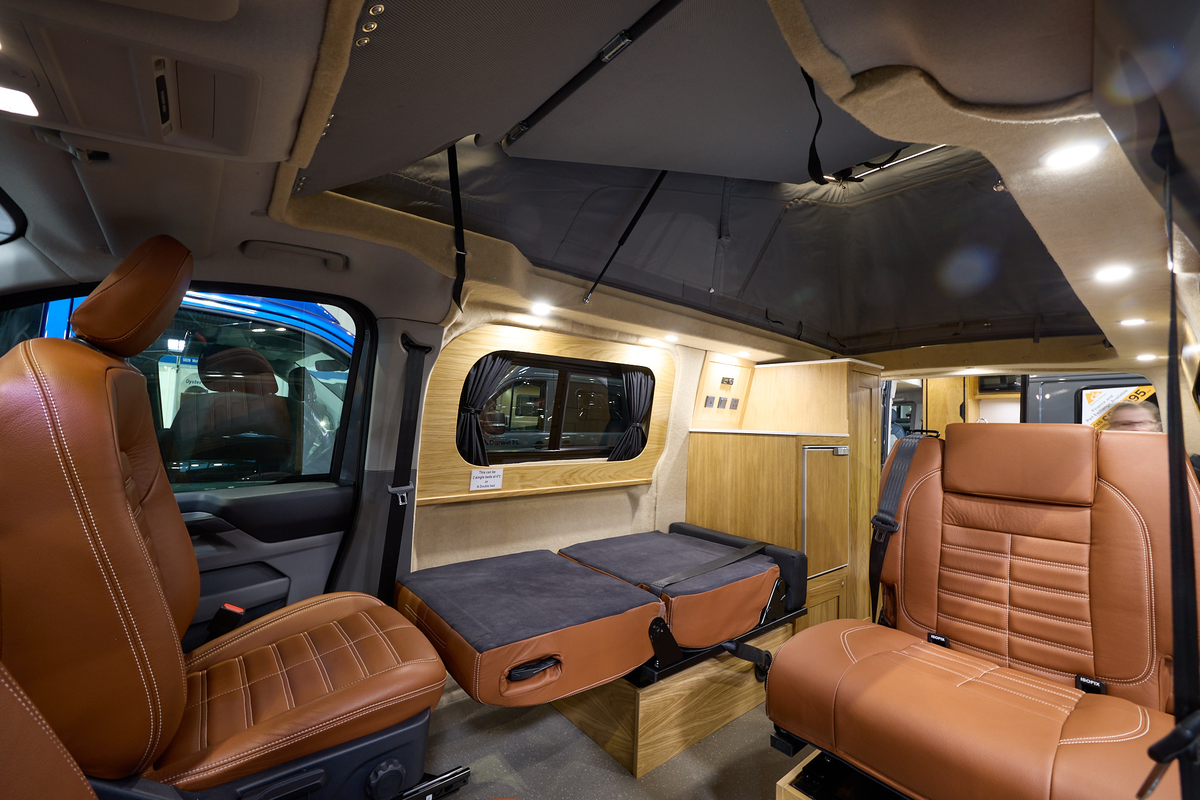 The Rolling Homes Camper Ltd Stanley has dark grey flooring.  The upholstery is brown leather.  The cabinets are wooden.  The rising roof is fully extended.  One of the passenger seats is folded down.