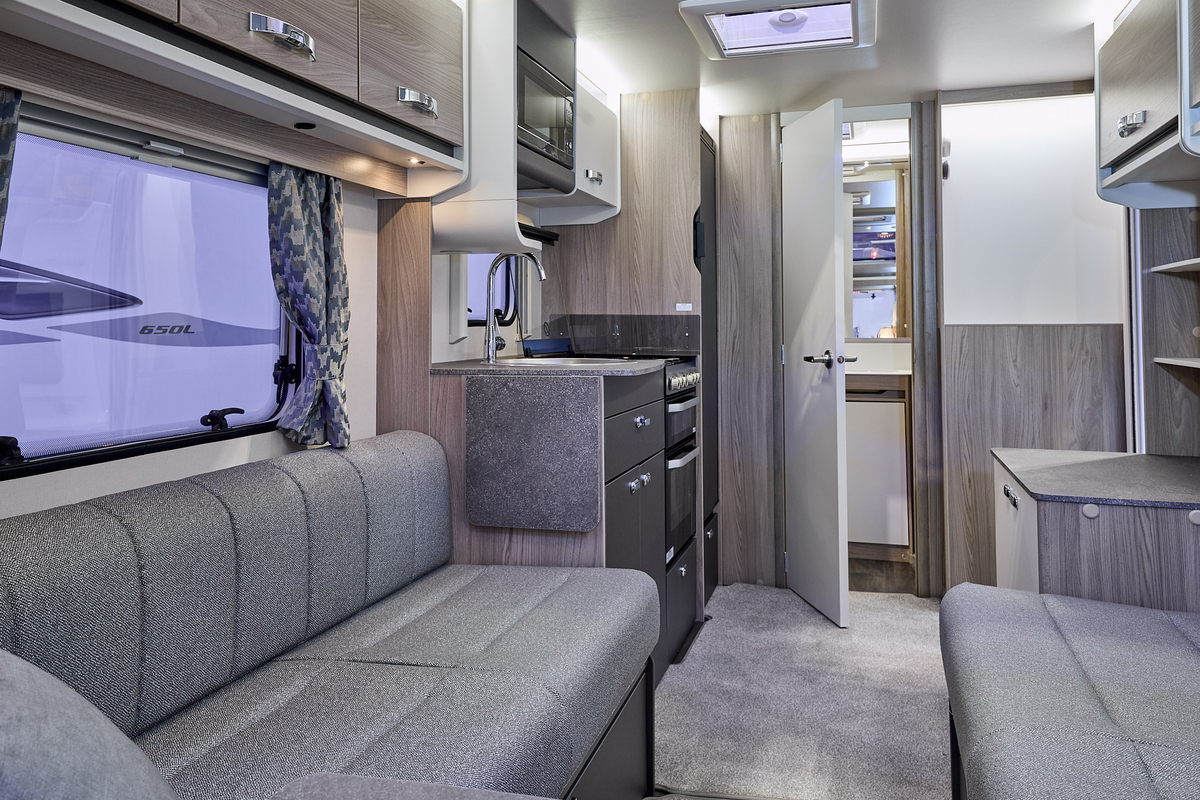 Swift Sprite Alpine 2’s interior has grey upholstery with wooden furniture.  The kitchen is in the middle and its furniture has dark doors. .  The rear door is open revealing the washroom