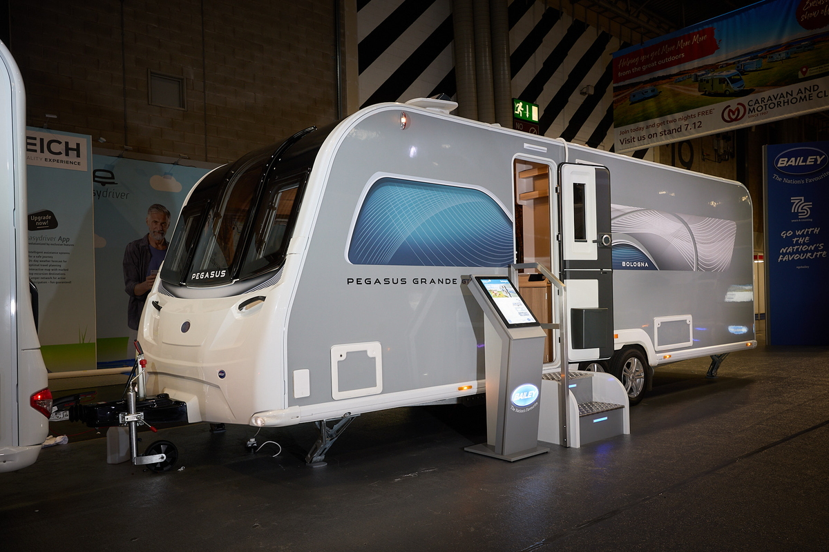 Bailey Pegasus Grande Bologna exterior’s, grey and white with a large single front window.  The entrance door is open and there are fixed metal steps into its interior.  There is an interactive stand next to the steps.