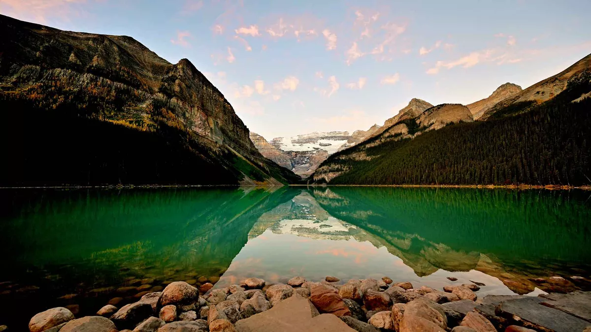 Looking out at ground level over Lake Louise in Banff National Park, Canada. The stones from the shoreline lead to the calm greenish water with the high mountains rising high in the distance on both sides. 
