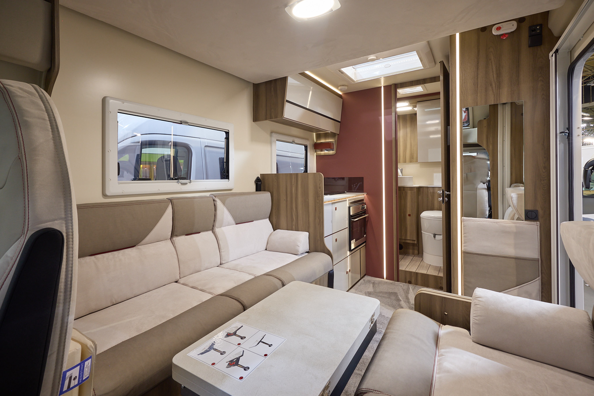 The Roller Team Kronos Compact 590 has grey carpet.  The upholstery is cream and mid brown.  The cabinets are cream with wooden surrounds and the interior door is a dark brown.  There is a step up to the rear washroom.