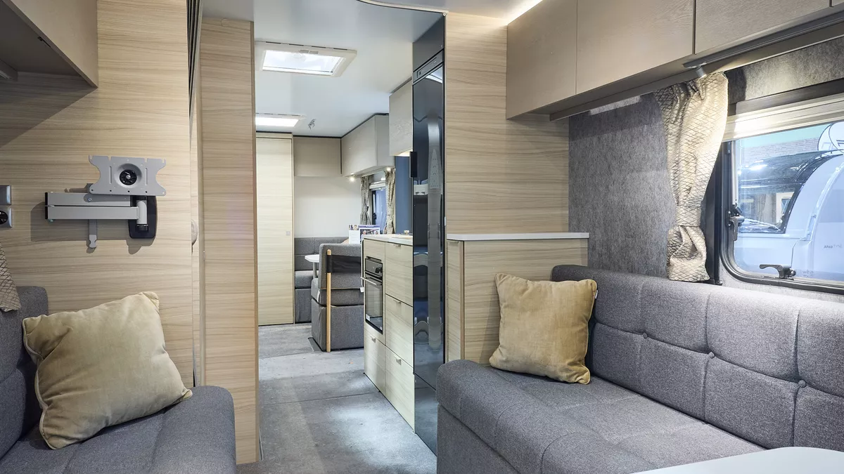 Interior of the Adria Altea Avon with grey sofas, cosy cushions and wood accents, illuminated by natural light through windows, offering a view outside.  Perfect for relaxation. and designed for comfort.