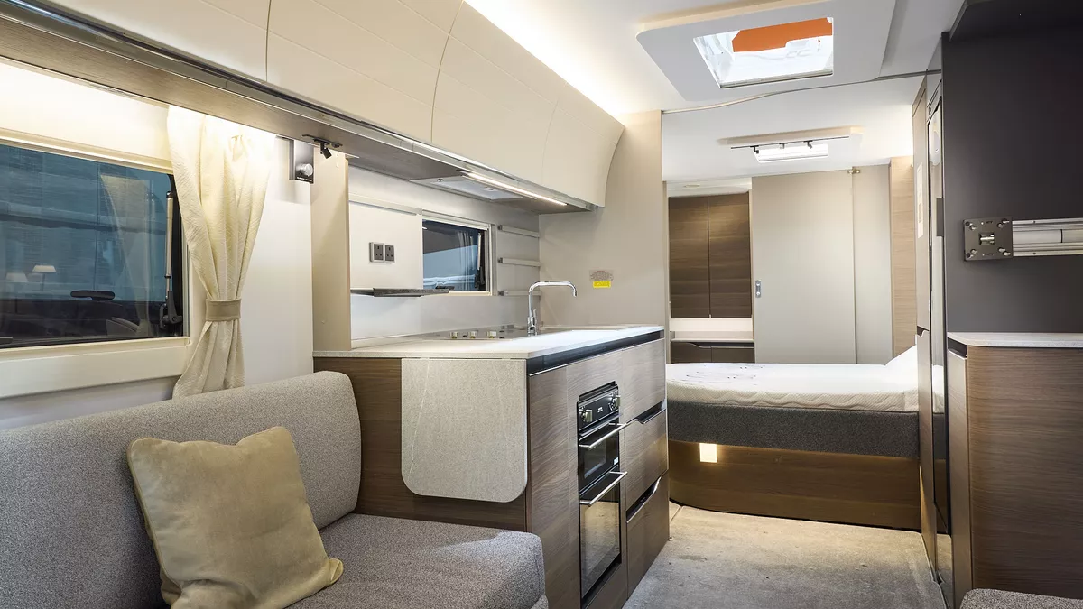 Interior of the Adria Alpina Mississippi with grey sofas, cosy cushions and wood accents, illuminated by natural light through windows and skylights.  There is a fixed island bed and the door at the rear leads to the washroom.