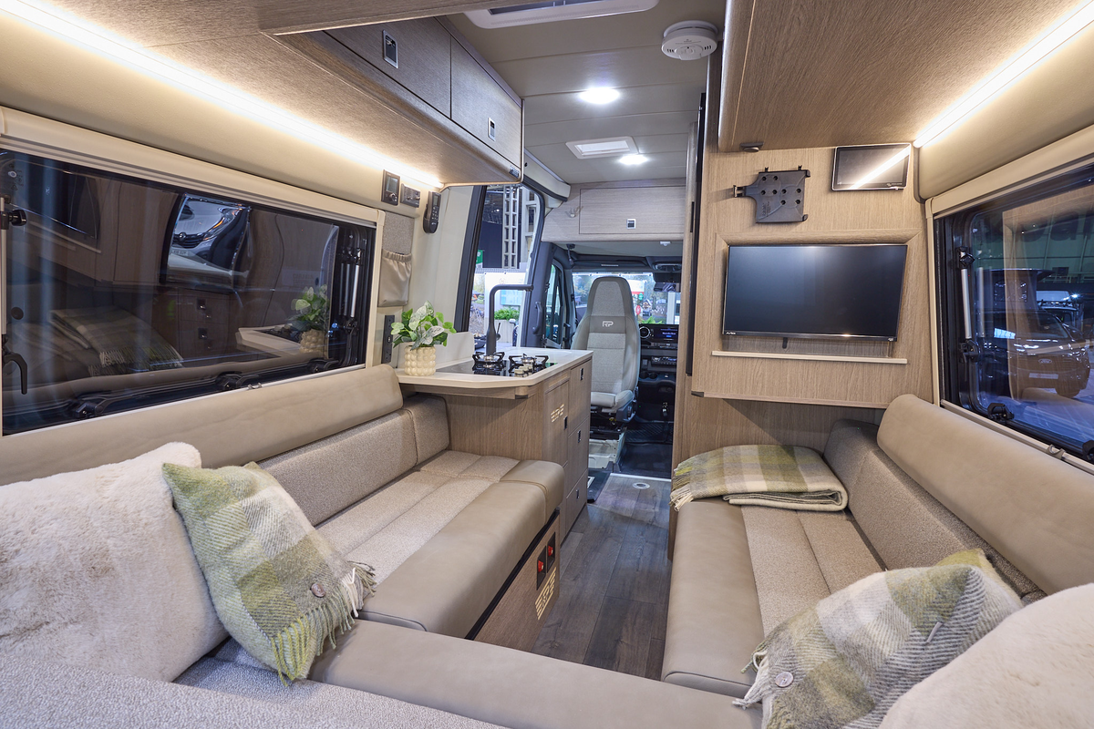 The RP Overland Rebel has wooden flooring.  The upholstery is cream with a matching pattern.  The cabinets are wooden.  There is a television on the right hand wall.  The passenger seat is facing into the vehicle.  There is a u shaped lounge.